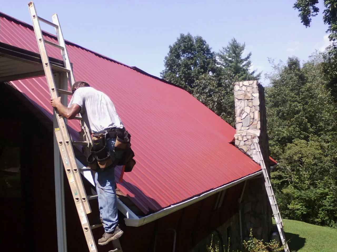 Roofer on a ladder beside a red metal roof and stone chimney — Soffit & Fascia Repair by Roofing Brothers in Tell City